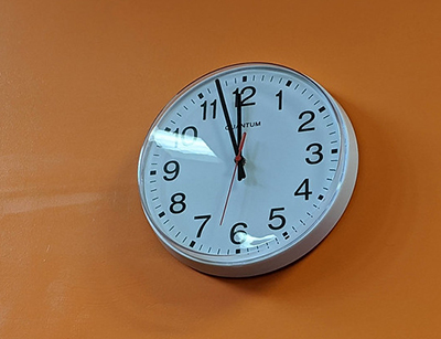 White analog clock showing the time 11:57, on an orange wall